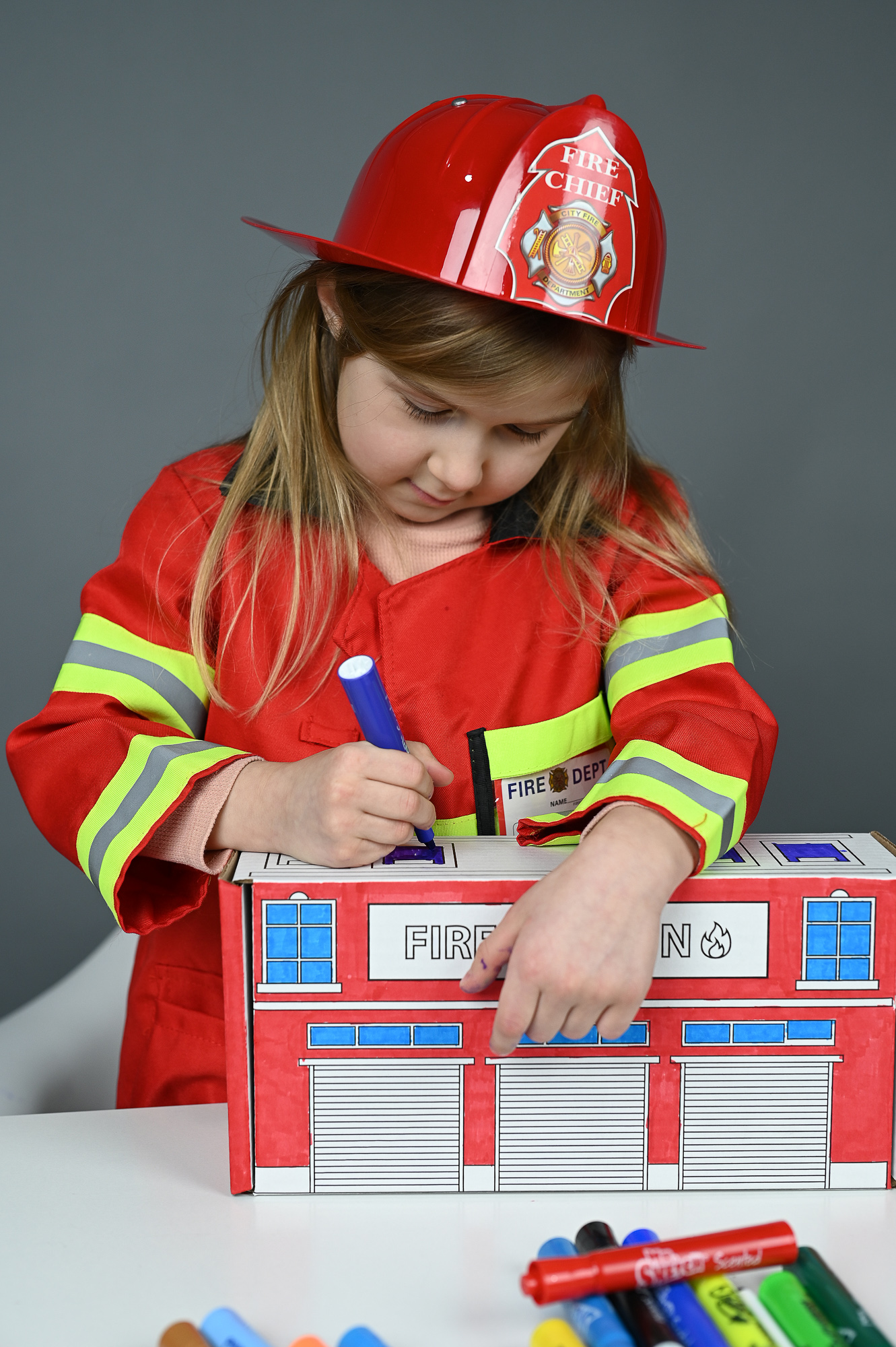 A child in a firefighter costume coloring the Fire Station box with a blue marker