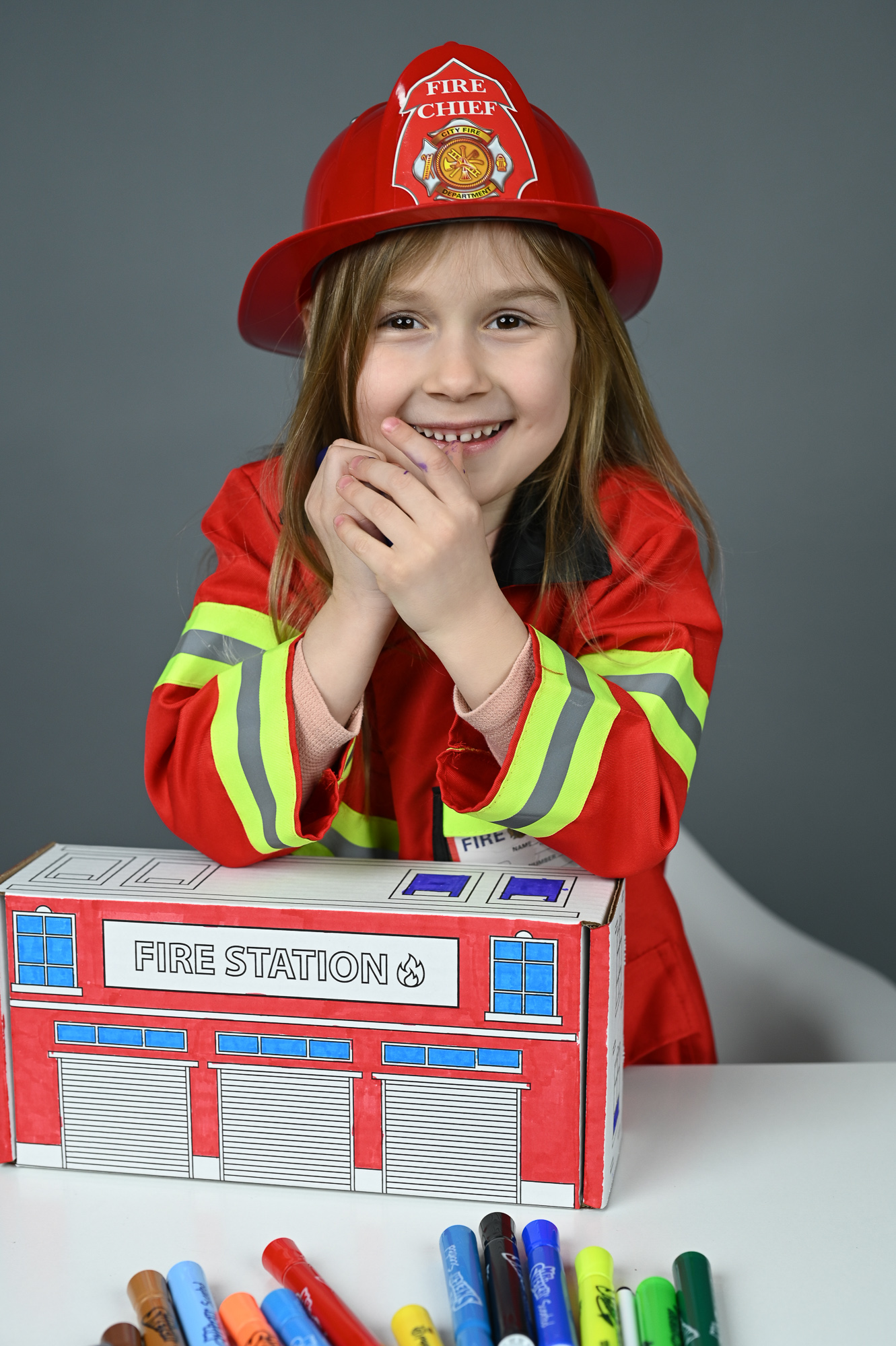 A smiling child in a fire chief helmet behind the finished red and blue Fire Station